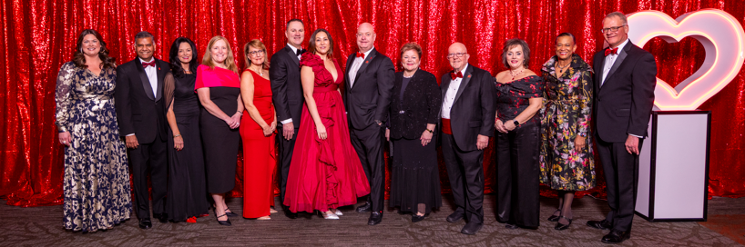 Group of men and women in formal wear standing next to a large lighted heart on a pedestal.